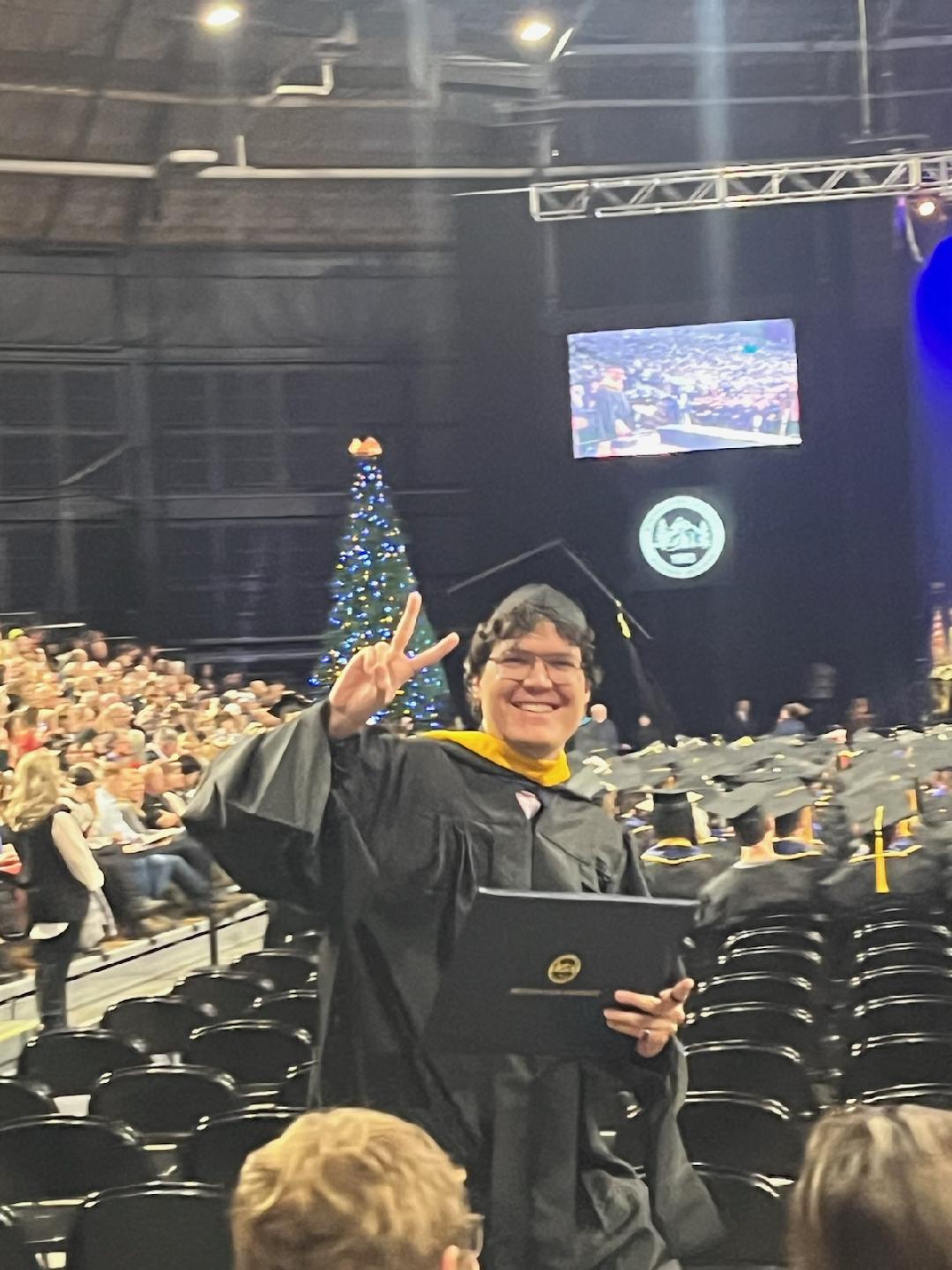 Garrett Figueroa wearing a graduation cap and gown, walking across a stage at Montana State University commencement ceremony, surrounded by other graduates and faculty in academic regalia, with a joyful and proud expression